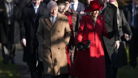PA Media King Charles dressed in a beige long coat walks along side Queen Camilla dressed in red