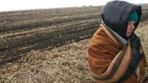 Svitlana Savinova Maria Savinova, wrapped up in a blanket and with a thick hood on her head, stands, eyes closed, in a barren field in Ukraine. There is snow on the horizon.