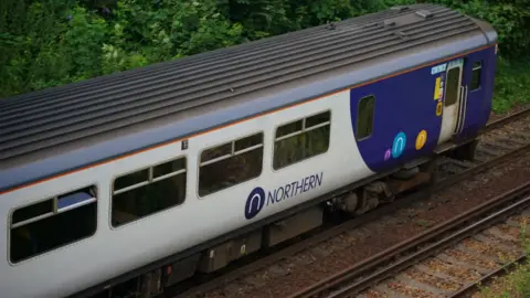 A blue-and-grey Northern train which is travelling along a two-track rail line. There are green trees and bushes to the side of the track.