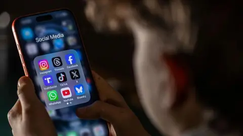 A child, who is out of focus, looks at a phone held in both hands. On the screen is a folder labelled 'social media' with app icons for Instagram, Threads, Facebook, TruthSocial, TikTok, X, WhatsApp, YouTube and Bluesky.