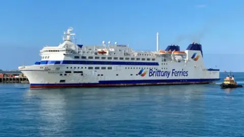 A Brittany Ferry sails toward the left of the screen. It is white with two blue stripes and blue writing. There is an escort boat to the rear left of it. 