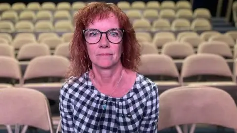 Sharon Catchpole, in a black and white gingham shirt, sits in the auditorium at the theatre, looking solemnly at the camera. She has red curly hair and fashionable black glasses