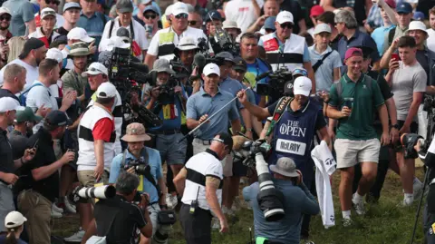Reuters Rory McIlroy carries his golf club as he walks through a crowd of photographers and camera operators. They are surrounded by scores of fans on a golf green. They are on the 17th hole at Royal Portrush Golf Club, Northern Ireland at the 2025 Open.