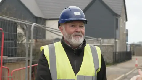 A man in a high viz jacket and a hard hat stands in front of a new housing development. He has a trimmed white beard.