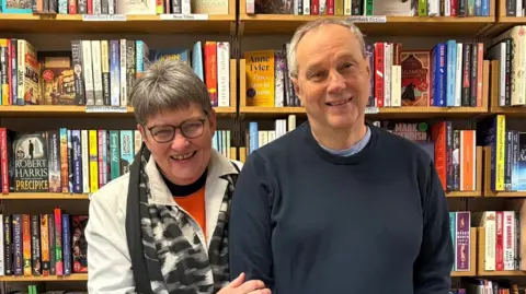 Brendon Books A woman and man stand next to each other looking at the camera smiling, arm in arm. Behind them are rows of books on shelves. 