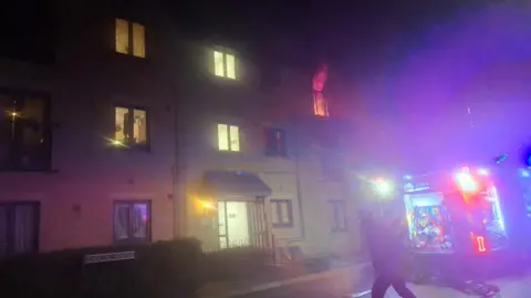 Chloe Raeburn A block of flats where fire is billowing out of one window. There is a fire engine at the scene and it is night time.