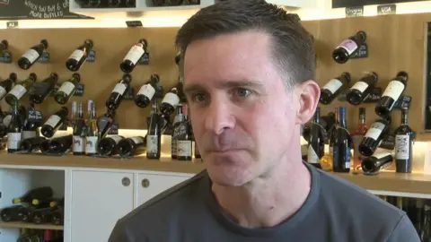 The picture shows a close-up of an adult indoors, framed from the chest upwards, with shelves of wine bottles filling the background. The person is wearing a plain, dark short‑sleeved top and is turned slightly to one side, looking just off camera. 