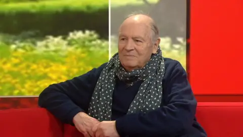 Charles has grey hair and is sitting on a red couch. He is wearing a dark blue jumper and a blue and white polka dot scarf. His hands are held together in front of him.