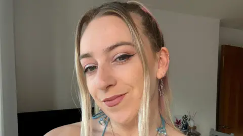 Abi White Abi smiles at the camera, her long blonde hair is pulled back from her face with strands at the front. She wears silver earrings and sparkly eyeshadow on her eyes.