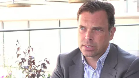 A close-up shot of Ken Skates being interviewed. He is wearing a grey suit jacket over a light blue and white striped shirt with an open collar and the background is very bright.