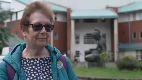 Lesley Mahmood has short brown hair and is wearing sunglasses and a green coat. The straps of a purple rucksack are showing over her coat. She is wearing a navy top with white flowers on it. She is standing outside the entrance to Liverpool's Women's Hospital. She is smiling.
