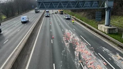 A motorway viewed from the bridge. Traffic officers have closed some of the lanes and a load of offal can be seen spread across several carriageways.