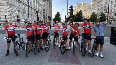 Ten members of Braveheart Bicycle Club, who are wearing helmets and cycling clothing, are standing next to their bikes with one hand in the air as they pose for a photograph in Liverpool. 