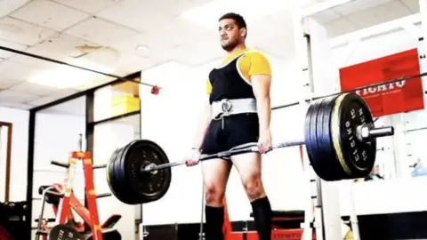 Bally Cheema A man is wearing a yellow top and a black unitard. He is holding a barbell with lots of weights.