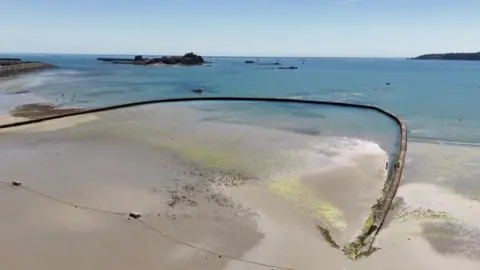 A drone shot looks down on a beach. A large, arcing concrete wall - which is supposed to hold water - starts on the sand and curves into and back out of sea. On the sand, a long yellow rope with several buoys lies along the sand on the entrance to the structure. The sea is turquoise under the blue sky, with a castle in the sea in the background.
