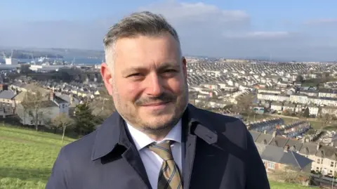 A man with a light beard and short grey hair looking at the camera in a navy jacket wearing a white shirt and pale brown coloured tie. There is a view of rooftops in a town behind him.