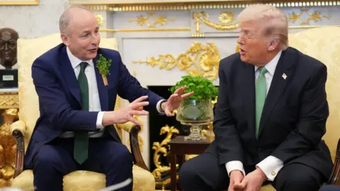 Martin and Trump are sitting next to each other in a room with a fire place, a bust of a head and a glass vase. Martin's hands are up as he talks. 