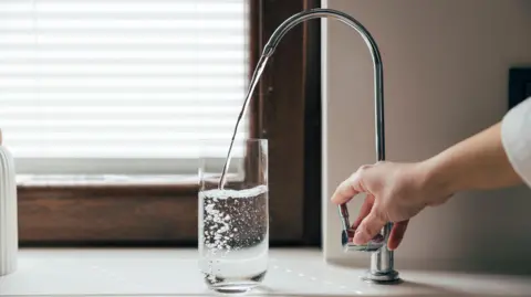 A hand turning a kitchen tap from which water is flowing into a glass