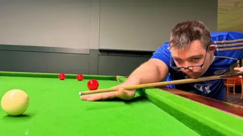 Dylan leaning low over a snooker table, aiming a cue stick at a white ball in the foreground, with three red balls positioned further behind on the green felt table. Dylan is wearing a blue sports t-shirt and black-framed specs low on his nose. He is in a brightly lit indoor venue.