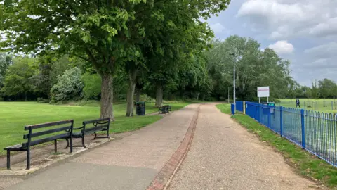 Shariqua Ahmed/BBC A brown walkway between two green spaces - lined with trees on side and a blue fence on the other side.