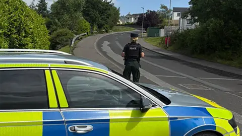 A police officer is standing in front of a police car. They are wearing full uniform including a hat. The background is a black road, footpath and crash barrier. There are houses and trees visible. 