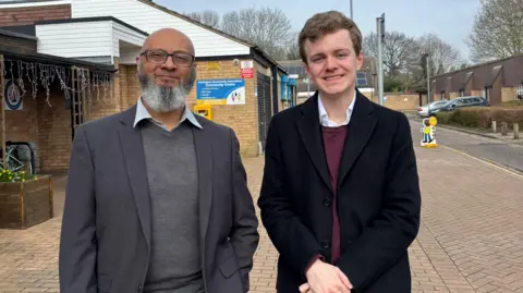 Peterborough City Council Ejaz, a man, wearing a grey blazer and grey cardigan, standing next to Sam Carling, a man in a black coat, smiling for the camera.