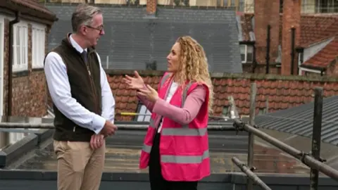 White and McCallion standing on what appears to be a rooftop or elevated outdoor work area. White is wearing a bright pink high-visibility vest with reflective silver stripes over a light pink jacket and dark trousers. McCallion is wearing a white shirt, a dark sleeveless fleece vest, beige trousers, and glasses.