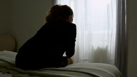 A generic picture of a woman sitting on a bed in a hotel room. She is still away from the camera and looking at a white curtain. 