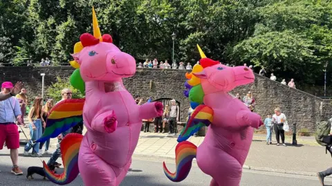 A parade with two people in large inflatable unicorn costumes, pink with rainbow manes, tails and wings
