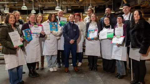 Blackpool Council 13 chefs in their white aprons line up alongside Blackpool Council catering manager Derek Wright. The majority of them are women, and they are holding up their certificates. 