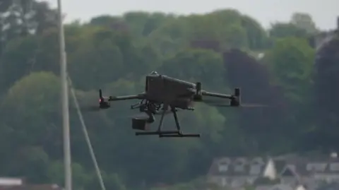 A black drone is in mid-air, with some buildings and trees in the background.