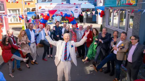 BBC 16 experts and presenters from Bargain Hunt form a semi-circle around another of their presenters. The presenter in the middle is a man in his 70s wearing a chalky white coloured suit and light blue shirt. The presenters surrounding him are all doing a kick in the air. They are all standing in a street with blue, white and red bunting under a happy 25th birthday Bargain Hunt banner. 