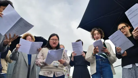 A choir stands in a circle, holding sheet music and singing.