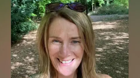 Laura Corby smiling in a selfie taken on a path through some trees on a sunny day.