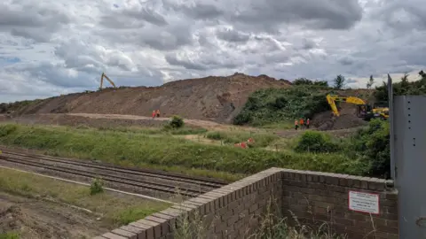 People in orange hi-vis workwear and diggers are working on a huge mound of soil many meters high. The mound is on the far side of a two-track railway line with patches of rough and sparse grass on either side. The sky above is overcast.