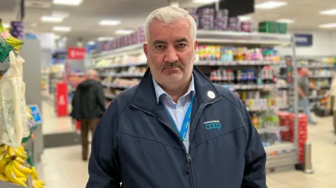 Mark Cox is standing in a store looking directly at the camera. He has short white hair and is wearing a Channel Islands Coop branded coat over a blue checked shirt and a lanyard is visible. To his right are bananas for sale and there are dairy aisles behind.
