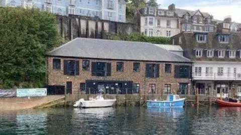 El edificio de Looe, en Cornualles, que alberga un centro bancario en su interior, se ve al otro lado del puerto, con barcos delante y otros edificios en lo alto detrás.