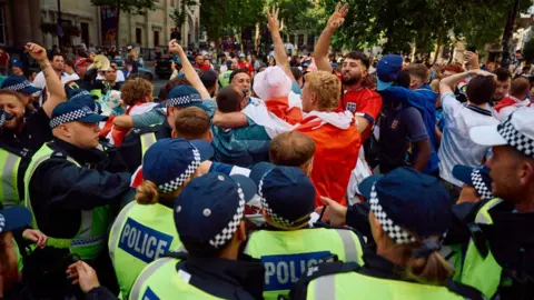 Police officers stand guard as England fans cheer for their team as they gather near Trafalgar Square in London ahead of the UEFA Euro 2024 final football match between Spain and England being played in Berlin on July 14, 2024. England are hoping to end their 58-year wait to win a men's major international tournament when they take on an outstanding Spain team in the final of Euro 2024.