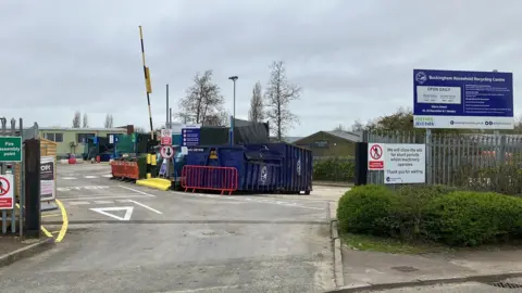 Buckinghamshire Council The entrance to a recycling centre, with a large sign to the right, blue containers at the entrance, a barrier up in the distance, and a green cabin on the left. There are several trees in the distance. There are lots of signs and a lamp post in the middle.