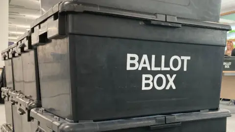 BBC A number of black plastic ballot boxes stacked on top of one another. The words 'BALLOT BOX' are printed in white on the side. 