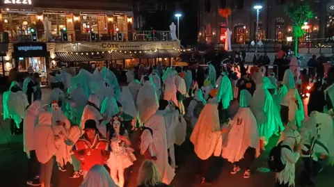 BBC A crowd of people dressed in white bed sheets and other Halloween costumes dance in a square. They are bathed in green and orange light.