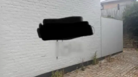 Peter Chaney Graffiti sprayed on a white wall of a housing building. There are words which have been blurred out with black and a clear blurring. There is a small brick yard in front of the side of the building. 