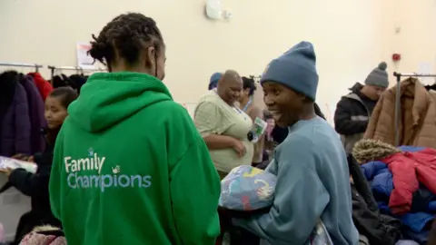 A woman wearing a green hoodie with the words Family Champions on the back is speaking to another lady wearing a light blue jumper and matching hat, who is smiling and holding a child's coat