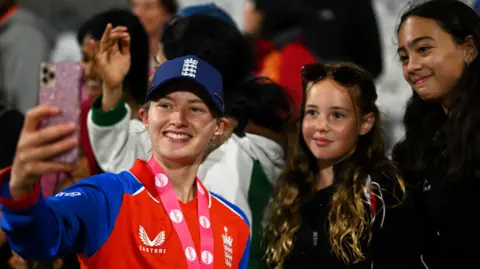 A mid-shot of Charlie standing in a crowd holding up a camera for a selfie and smiling along with two young female cricket fans