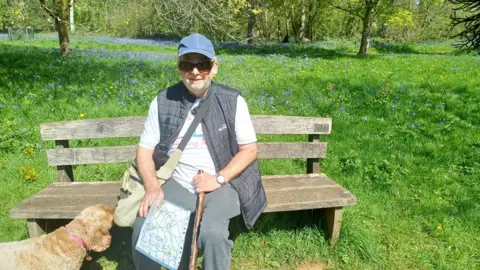 Roy Howard A man sitting on a bench on a sunny day. There is a cream-coloured dog sniffing the bag that is across the man's body. The man is holding a map and a walking stick and is wearing a blue cap, a bodywarmer, grey trousers and a white T-shirt.