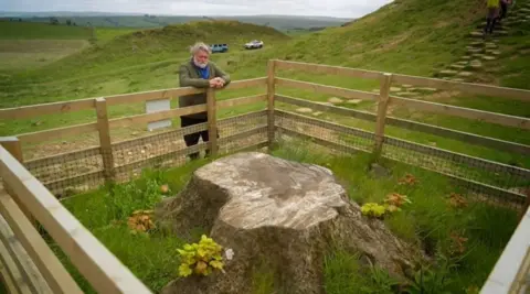 Si King is standing at the stump of Sycamore Gap, which has a fence around it. On one side there are the paving stones that make up Hadrian's Wall. He has grey hair and a beard. He is wearing a blue jumper and a jacket and black trousers. In the background you can see two cars parked on a grass track and green hills