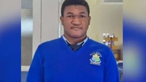 Handout Emmanuel Familola wearing his school uniform. He has short, dark hair and is wearing a bright blue jumper, a light blue shirt and a navy tie.