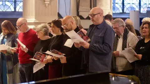 A crowd of people singing carols from sheets of paper. They range from some men with shaved heads and glasses nearer the camera to women with short grey hair or longer dark hair, all singing in a church in wooden pews.