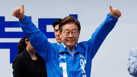 Lee Jae–myung in a blue tracksuit jacket and glasses lifts both hands into the air with thumbs raised