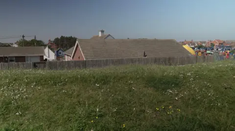 A large grass sea defence is in the foreground and behind it the rooftops of a number of low-lying bungalows, with the roofs almost level with the top of the grass bank.